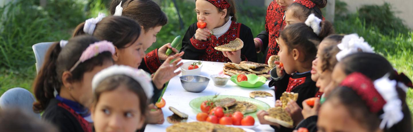 Children eating at a table outside with food.