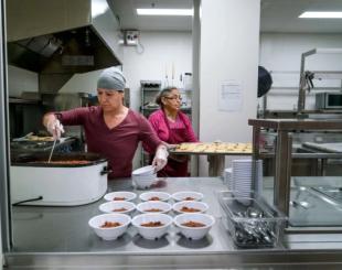 Two women in a commercial kitchen serve food into bowls.